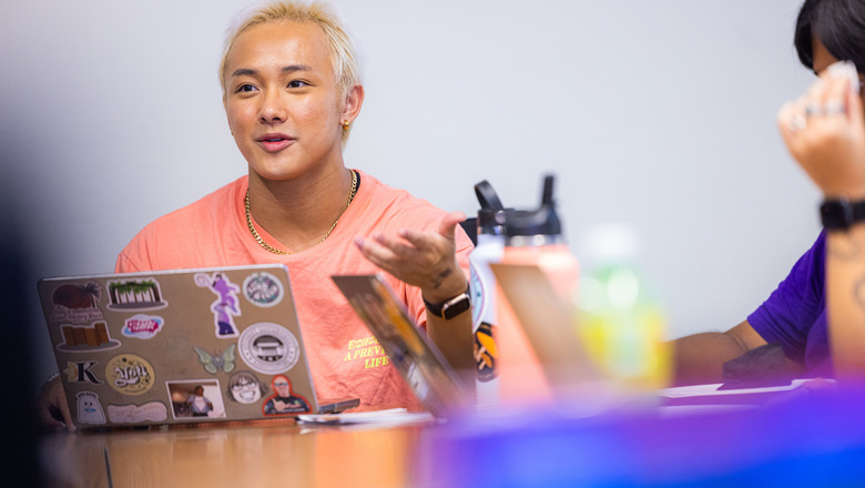 Students have a a discussion in class. There are laptops and water bottles with stickers on them on the desks.