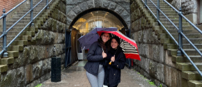 Two students stand in front of brick building holding umbrellas while studying abroad