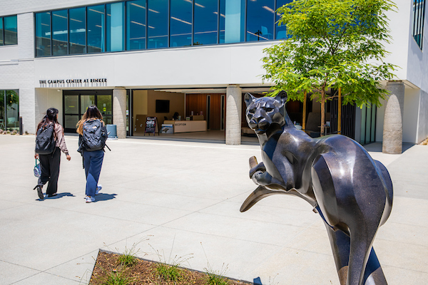 Two students walk outside on Rinker Campus. They are walking towards the entrance to a building, with a statue of a panther in the foreground.