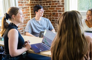 Students working on laptops at a desk in an office environment. They are having a discussion and smiling.