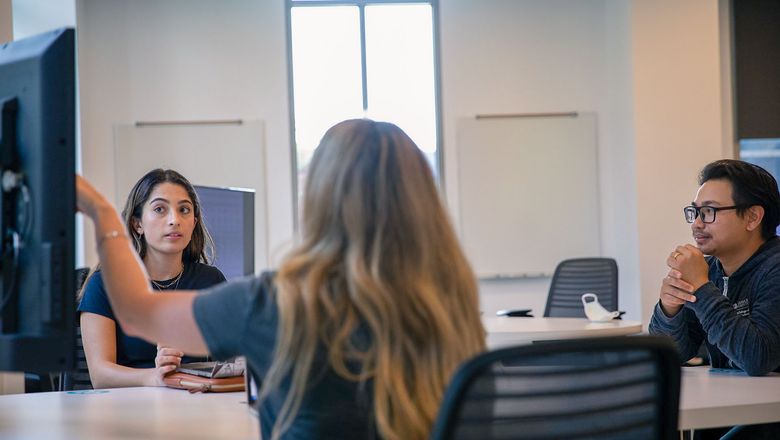 Three students engage in a discussion in front of a screen in a conference room.