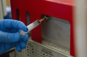 A blue-gloved hand inserts a syringe into a machine.