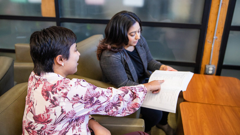 Two people sit next to each other in an office setting. One is holding a sheet of paper or binder while the other points to it.