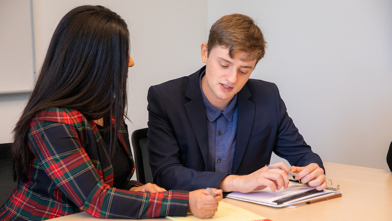 Two students look over and discuss notes at a desk. One of them is professionally dressed.