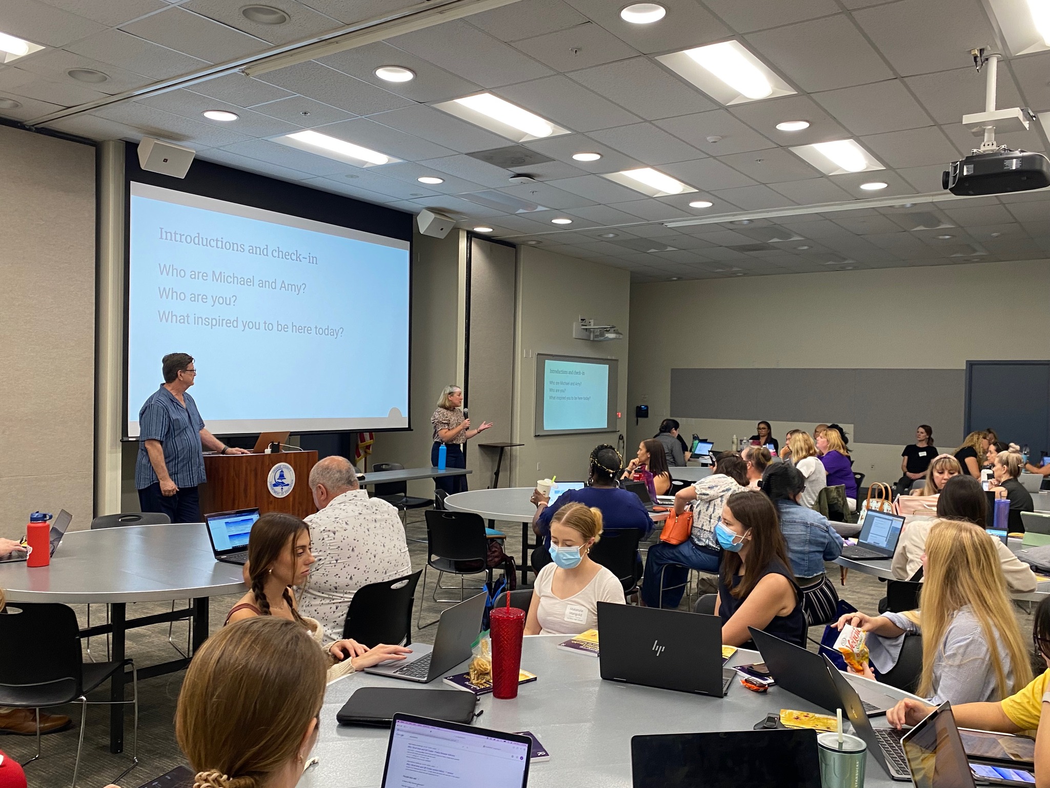 Dr. Ardell is presenting in front of a room of people sitting at tables, taking notes on laptops. Dr. Hass is standing next to the lectern watching Dr. Ardell present. A large screen for the slides is behind Drs. Ardell and Hass. 