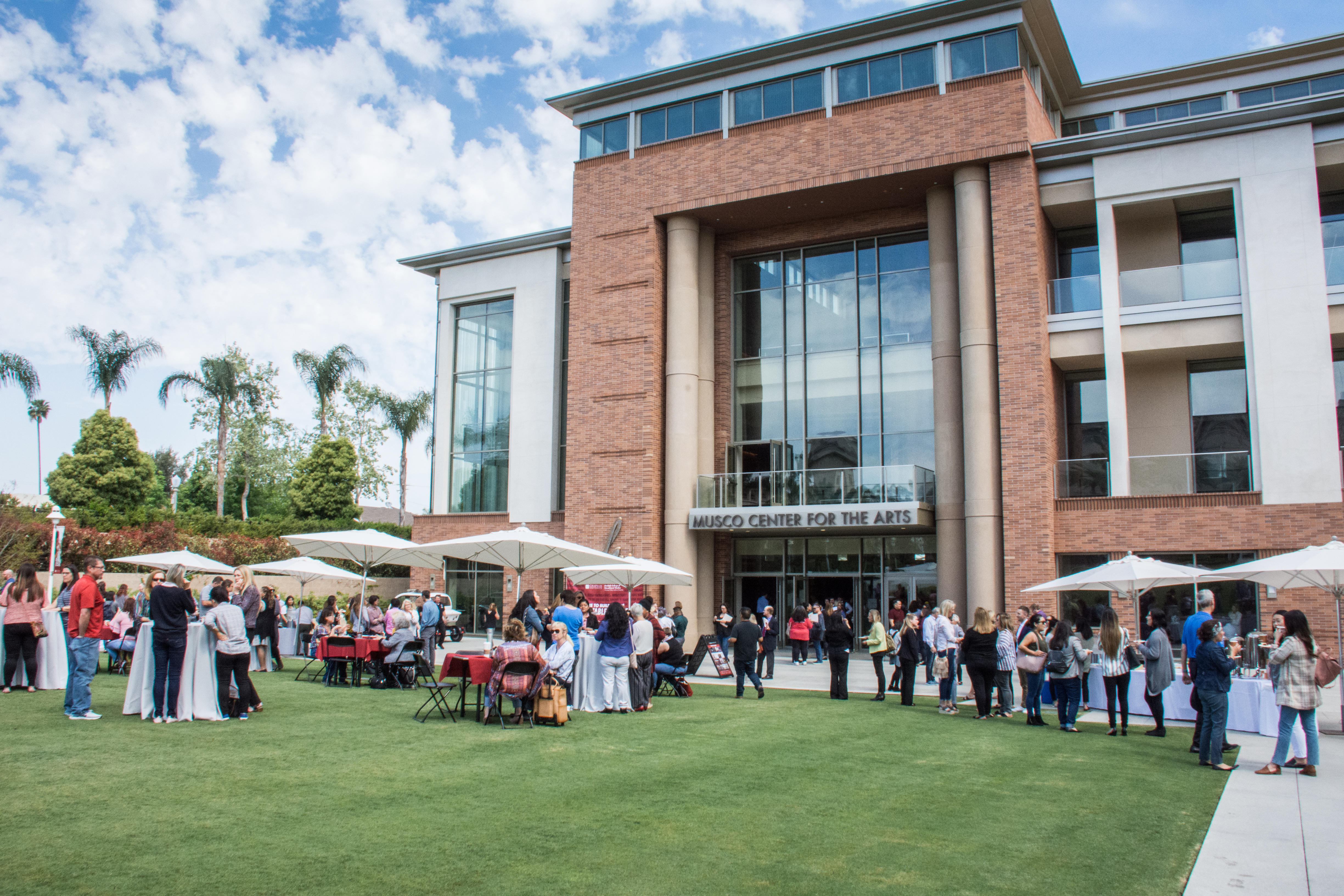 Exterior of Musco Center for the Arts. The Musco Center of the Arts is three story building with windows from first floor to third floor. In front of Musco is a large green lawn with tables set up on the lawn. Attendees of the Summit are standing around the tables talking and mingling on the lawn.