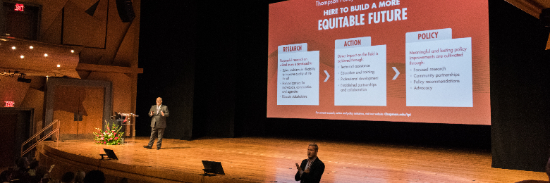 Dr. Don Cardinal is presenting on stage with a screen in the background that reads "How to Build a More Equitable Future: Research, Action, Policy. An ASL interpreter is standing in front of the stage.