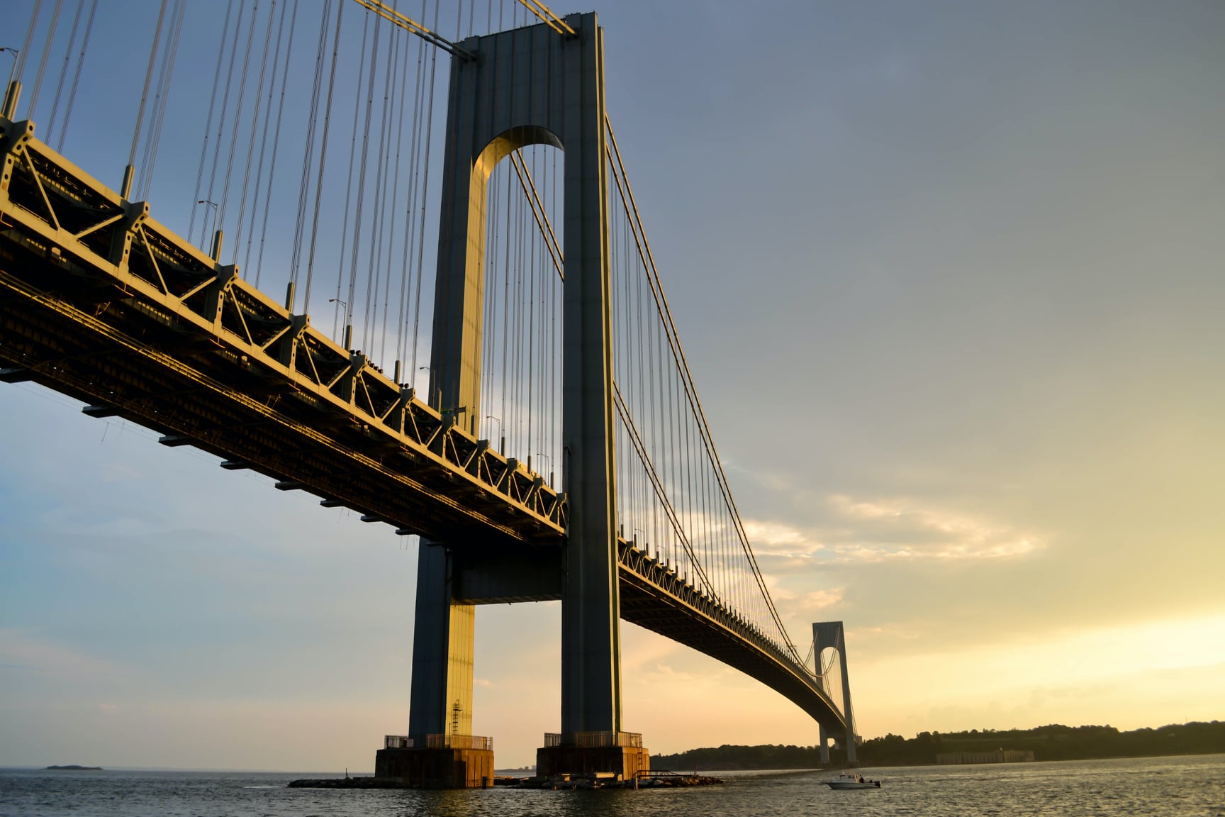 Image is of a large, metal bridge spanning a body of water to a distant land as the sun rises.