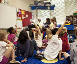 A classroom with students sitting on the floor facing a teacher who is holding a book - photo by CDC on Unsplash