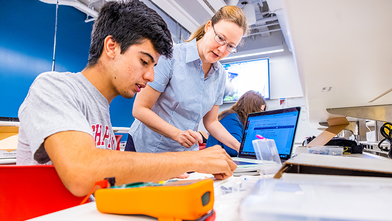 A student works on assembling a device in a class while a professor looks on.