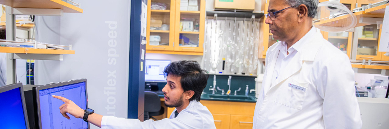 Student sits at desk in lab and points at computer screen while another person looks at screen behind student