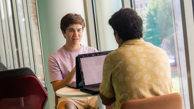 Two students have a discussion while sitting at a desk in a brightly lit study area. There are laptops on the desk.