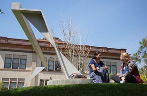 Two students converse while sitting on a lawn next to an abstract structure.